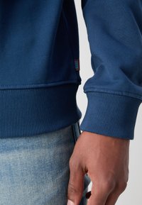 Felpa blu navy con polsini e orlo a costine. Presenta un'etichetta con logo sul lato, una texture liscia e un design aderente. Indossata con jeans azzurri.