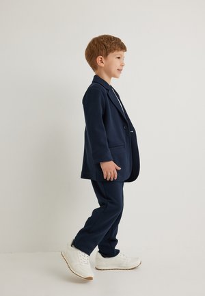 Chaqueta de traje azul marino con solapas de muesca y un solo botón, combinada con pantalones a juego y zapatillas blancas, con una textura suave.