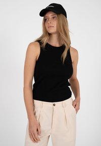 Femme aux longs cheveux blonds portant un haut noir sans manches, un pantalon beige et une casquette noire, regardant sur le côté avec les mains dans les poches.