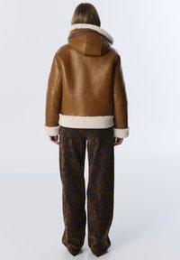 Bruine leren jas met een witte nepbont kraag en manchetten, gecombineerd met losse, gemusterde broeken in bruine en donkerblauwe tinten.