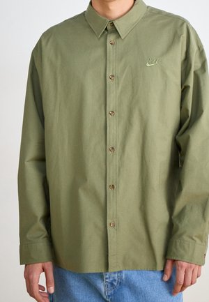 Mężczyzna ubrany w oliwkową koszulę na długi rękaw zapinaną na guziki z logo Nike na piersi oraz jasnoniebieskie dżinsy, stoi z rozluźnionymi rękami wzdłuż ciała.