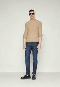 Μπεζ ριμπ τζίν γγλυνός, μπλε στενά τζιν, μαύρα παπούτσια και γυαλιά ηλίου; μοντέλο στέκεται μπροστά σε ουδέτερο φόντο.