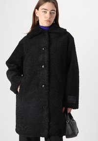 Cappotto nero e soffice realizzato in tessuto testurizzato; presenta un colletto, bottoni frontali e un'etichetta del marchio sulla manica. Design semplice con tasche.