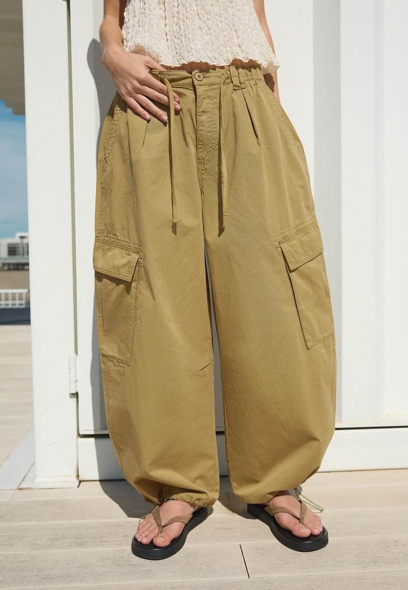 Personne portant un pantalon cargo ample kaki avec taille à cordon et poignets élastiques, un haut sans manches beige texturé et des tongs noires.