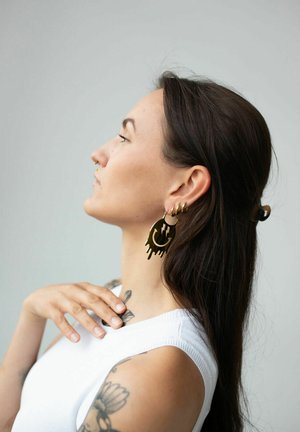 Gold-tone hoop earrings with abstract dripping design, worn by a model with long hair, featuring a white sleeveless top and visible tattoos.