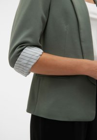 Marynarka w kolorze oliwkowej zieleni o wyraźnie zarysowanym kształcie, z podwiniętymi rękawami odsłaniającymi biało-zieloną, paskowaną tkaninę. Miękka faktura, minimalistyczny design.