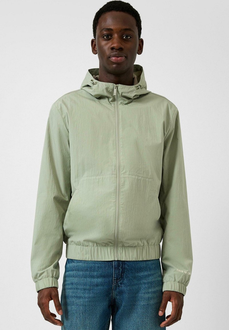 Jeune homme portant une veste à capuche vert clair avec fermeture éclair, poignets et ourlet élastiqués, associée à un jean bleu, faisant face à l'avant.