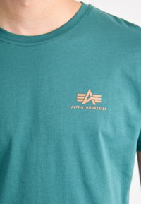 Turkizna bombažna majica s krožno izrezano ovratnikom, ki ima majhen oranžen logo in grafični dizajn "Alpha Industries" na levem prsnem delu.