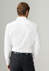 Chemise blanche à manches longues avec un col classique, vue de dos, associée à un pantalon noir et une ceinture noire brillante.