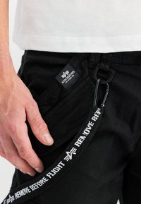 Schwarze Cargo-Hosen mit einem auffälligen "REMOVE BEFORE FLIGHT"-Riemen und einem Etikett von Alpha Industries, aus einem glatten, strapazierfähigen Stoff.
