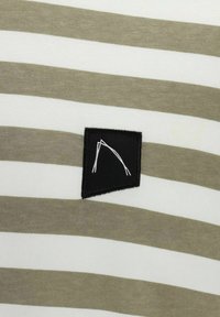 Juostuota trumpomis rankovėmis marškiniai, kuriuose pakaitomis yra alyvuogių žalios ir baltos juostos, su juodu kvadratiniu pleistru, ant kurio pavaizduota balta geometrinė forma.