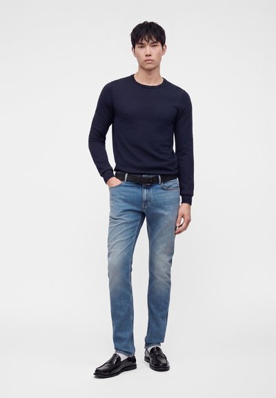 Jeune homme debout avec une main dans la poche, portant une chemise noire à manches longues, un jean bleu, une ceinture noire et des chaussures habillées noires.