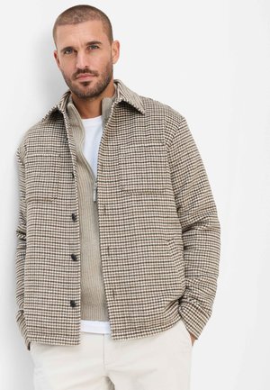 Mand med kort hår og skæg iført beige ternet jakke over beige lynlåstrøje og hvide bukser, stående mod en hvid baggrund.