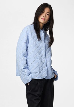 Junge Frau trägt eine hellblaue gesteppte Jacke mit diagonalen Mustern und großen Taschen, kombiniert mit schwarzen Hosen mit Nadelstreifen.