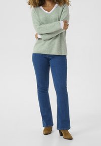 Jasnozielony sweter o fakturze z białymi wykończeniami, w połączeniu z niebieskimi spodniami dzwonami i brązowymi botkami na obcasie. Modelka pozuje z rękami skrzyżowanymi.