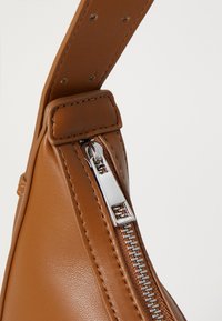 Coin de sac en cuir marron avec une fermeture éclair en métal et des coutures, doté d'une bandoulière robuste et d'une texture lisse.