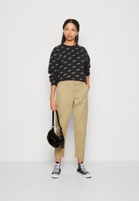 Felpa nera con logo bianco ripetuto, chinos beige, sneaker nere e piccola borsa nera. Modella in piedi con le mani sui fianchi.