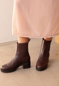 Bottes en cuir marron à cheville avec un bout arrondi, des panneaux latéraux texturés, des talons bas et des semelles à crampons, associées à une jupe rose clair.