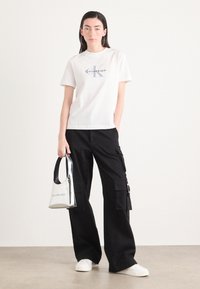 Camiseta de algodón blanca con logo, pantalones cargo negros con bolsillos laterales, bolso plateado y zapatillas blancas sobre un suelo beige.
