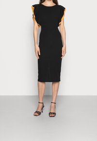 Robe midi ajustée noire avec des manches volantées jaunes en accent. Texture lisse. Associée à des talons ouverts noirs à brides.