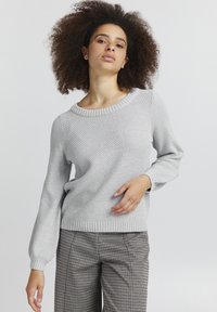 Helles graues Strickpullover mit einem strukturierten Muster, weitem Ausschnitt und langen Ärmeln. Kombiniert mit karierten Hosen, die eine bequeme Passform betonen.