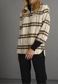 Camicia a quadri beige con righe orizzontali marroni e blu, due tasche sul petto, bottoni e vestibilità rilassata. I pantaloni neri completano l'outfit.