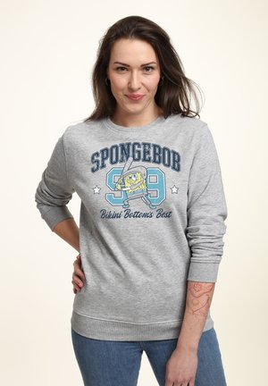 Frau mit langen dunklen Haaren, die einen grauen Pullover mit SpongeBob spielt Baseball und dem Text „Bikini Bottom's Best“ trägt.