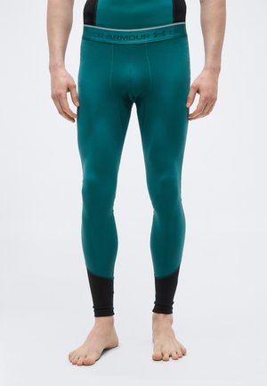 Mann trägt türkise Under Armour Kompressionsleggings mit schwarzen Knöchelpaneelen, steht barfuß vor einem schlichten Hintergrund.