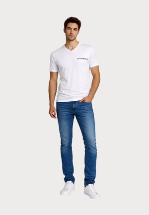 Mladý muž oblečený v bielom tričku s výstrihom do V značky Emporio Armani, modrých džínsach a bielych teniskách, stojaci s jednou rukou v vrecku.