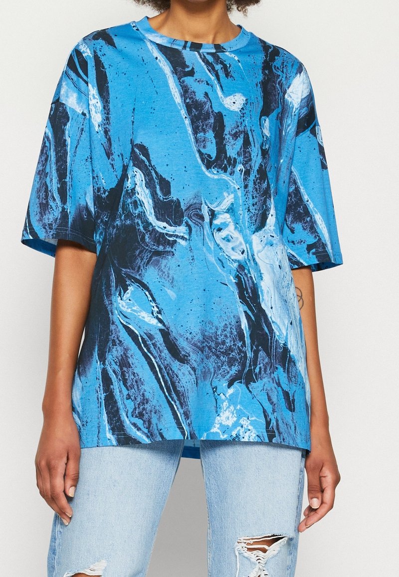 Personne portant un T-shirt oversize à motifs abstraits bleus et noirs avec un jean déchiré bleu clair, debout devant un fond uni.