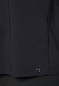 Haut en tissu noir avec une texture lisse, un ourlet incurvé et un petit accent métallique circulaire sur la couture latérale inférieure.