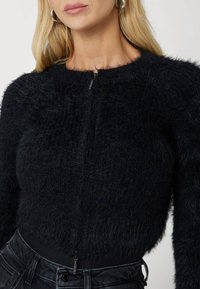 Black, fluffy knit cardigan with long sleeves and a zip closure, featuring a textured pattern and a fitted silhouette.