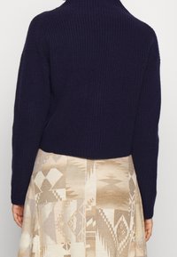 Marineblauwe, geribbelde gebreide trui met een hoge kraag, kort model. Gecombineerd met een beige rok met geometrische patronen.