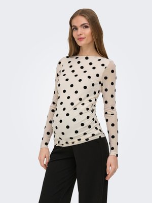 Woman wearing a beige long-sleeve top with black polka dots and black pants, standing with a slight smile against a plain background.
