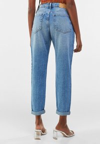 Světlé modré džíny s vysokým pasem a ohrnutými nohavicemi, dvěma zadními kapsami a texturovaným povrchem. Spárovány s sandály na průhledném podpatku.