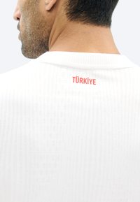 Άνδρας με κοντά σκουρόχρωμα μαλλιά που φοράει λευκό υφασμάτινο πουκάμισο με την επιγραφή "TÜRKİYE" σε κόκκινο στο πάνω μέρος της πλάτης.