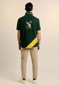 Camiseta polo verde con detalles amarillos y logotipo. Presenta un cuello y mangas cortas. Combinada con pantalones beige claros y zapatillas blancas.