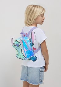 Fille jeune portant un sac à dos holographique avec un design de personnage de dessin animé, un t-shirt blanc et une jupe en jean, se tenant de profil devant un fond uni.