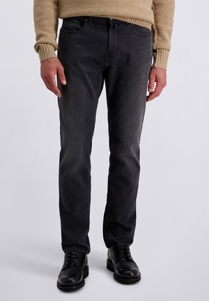 Mann in schwarzen, engen Jeans, schwarzen, ledernen Schnürschuhen und einem beigefarbenen Strickpullover vor einem einfarbigen Hintergrund.