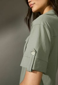 Camicia a maniche corte di colore verde oliva, realizzata in tessuto morbido, con dettagli in bottone e cuciture sul polsino per un aspetto strutturato.
