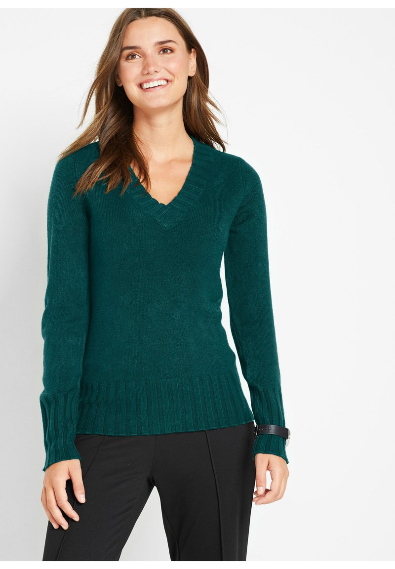 Maglione teal a V, realizzato in un materiale morbido, con polsini e orlo a coste. Indossato con pantaloni neri, offre una vestibilità confortevole.