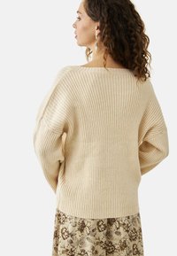 Jersey de punto beige con textura de acanalado, mangas anchas y un ajuste holgado, usado sobre una falda floral. Cuello redondo en la parte posterior.