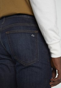 Pantalones vaqueros de mezclilla azul oscuro con costuras amarillas en contraste, que presentan un bolsillo trasero con un pequeño logo blanco y una textura suave.