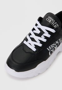 Črne usnjene superge z belimi vezalkami, ki imajo blagovno znamko "Versace Jeans Couture". Debel bel podplat z teksturiranim gumijastim oprijemom.