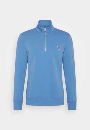 Mann trägt einen blauen Sweatshirt mit Viertel-Reißverschluss und passende Shorts mit kleinem Zebra-Logo, lehnt an einem Holzrahmen vor beigem Hintergrund.