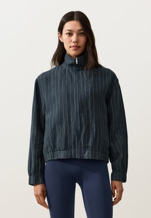 Hosszú, sötét hajú nő sötét tűcsíkos cipzáros dzsekit és magas nyakú, tengerészkék leggingset visel, és előre néz, egyszerű háttér előtt.