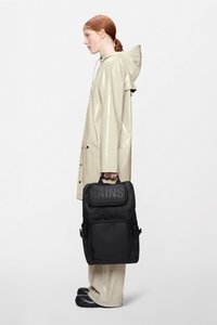 Μαύρο αδιάβροχο σακίδιο με μπροστινή τσέπη, με τη μάρκα "RAINS", κρατιέται από ένα άτομο με ανοιχτό μπεζ γυαλιστερό αδιάβροχο.