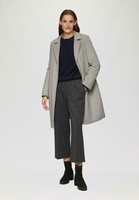 Grijze wollen jas, marineblauwe geribbelde trui, gestructureerde donkergrijze wijde broek en zwarte enkelboots. Model staat met de handen in de zakken.