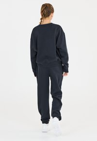 Marinfärgad sweatshirt och joggers i mjukt material, med en avslappnad passform och ribbade muddar. Modellen bär vita träningsskor, sedd bakifrån.