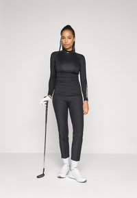 Mulher vestindo uma roupa atlética preta, segurando um taco de golfe, usando luva branca, meias brancas e ténis brancos, de pé contra um fundo simples.